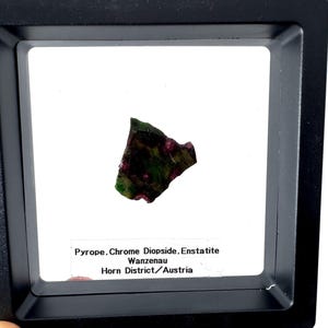 Peut inclure: Une petite pierre précieuse de forme irrégulière, avec un mélange de teintes vertes et rouges, présentée dans un coffret noir. Le texte sur l'étiquette indique : Pyrope, Chrome Diopside, Enstatite, Wanzenau, Horn District/Austria.