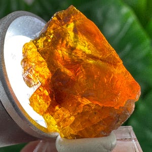 Peut inclure: Une pierre précieuse orange brute et translucide avec une texture cristalline irrégulière. La pierre est éclairée par l'arrière par une source lumineuse circulaire, soulignant ses teintes ambrées et dorées, ses bords tranchants et ses fissures.