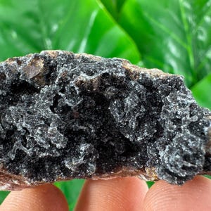 May include: A close-up of a dark, textured mineral specimen. The rock has a rough, uneven surface with a mix of dark gray and brown tones. The mineral appears to have a crystalline structure, with small, sparkling inclusions.