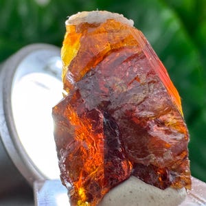Peut inclure: Un spécimen minéral brut et translucide présentant des teintes ambre, orange et brun rougeâtre profond. Le cristal possède une surface dentelée et multifacette avec des fractures internes visibles et un aspect lumineux et éclatant.