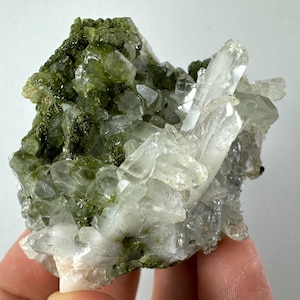 May include: A cluster of clear quartz crystals with green epidote inclusions. The crystals are clustered together in a natural formation.