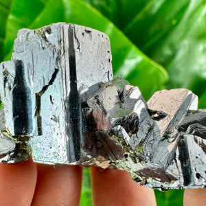 May include: A cluster of metallic gray, silver, and black crystals with a sharp, geometric shape. The crystals are arranged in a cluster, with some crystals overlapping others. The crystals have a shiny, reflective surface.