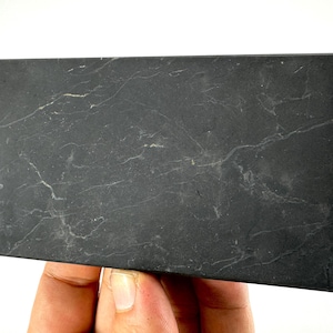 May include: A rectangular, dark gray stone with white veining. The stone is held in a hand and appears to be a decorative or functional item. The surface has a natural, textured appearance.