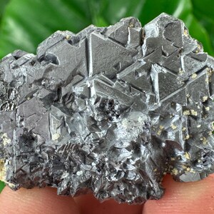May include: A raw galena mineral specimen featuring a cluster of metallic, dark gray, cubic crystals with sharp, geometric edges and a reflective, lead-like luster. The crystalline structure displays distinct triangular and square facets.