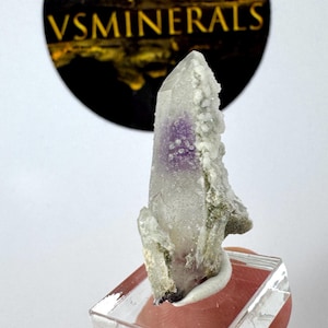 May include: A clear quartz crystal with a vibrant purple center and white, textured edges. The crystal is mounted on a clear, rectangular base. The logo "VSMINERALS" is visible in the background.
