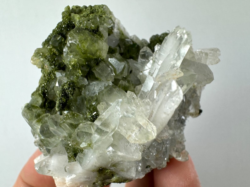 May include: A cluster of clear quartz crystals with green epidote inclusions. The quartz crystals are sharp and well-formed, and the epidote is a deep green color.