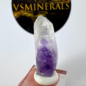 Può includere: Una punta di cristallo di ametista naturale, che mostra una sfumatura di colori dal trasparente al viola intenso. Il cristallo è montato su una piccola base circolare bianca. Lo sfondo include un logo con la scritta "VSMINERALS".