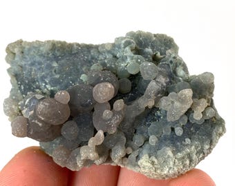 May include: A close-up of a blue-grey mineral specimen with a bubbly, botryoidal structure. The surface is covered in small, rounded formations, with a mix of light and dark grey hues. The overall texture appears rough and uneven.