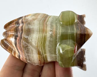 May include: A carved frog figurine made of layered onyx stone. The frog is primarily green, brown, and white with a polished finish. The figurine is held in a hand, showcasing the natural stone's patterns and colors.
