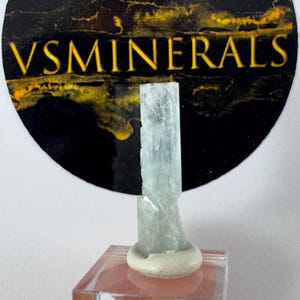 May include: A pale blue aquamarine crystal specimen. The crystal is mounted on a clear acrylic stand with a white base. The background features a black and gold circular logo with the text "VSMINERALS".