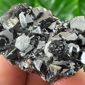 May include: A cluster of dark, metallic-looking crystals with sharp, geometric shapes. The crystals are primarily black with some areas reflecting light, creating a shiny appearance. The overall structure is rough and irregular, showcasing natural mineral formations.