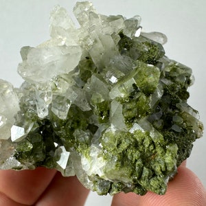 May include: A cluster of clear quartz crystals with green inclusions. The crystals are clustered together in a rough, natural formation.