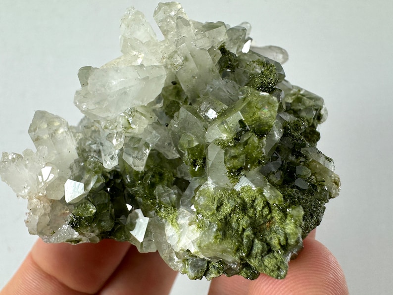 May include: A cluster of clear quartz crystals with green inclusions. The crystals are clustered together in a rough, natural formation.