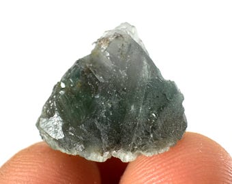 May include: A rough-cut, translucent mineral specimen with shades of green and gray. The crystal is held between two fingers, showcasing its natural, irregular shape and texture. The background is plain white.