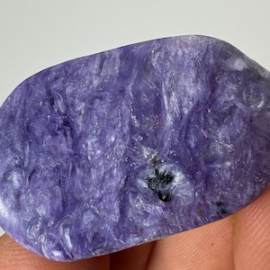 Puede incluir: Una piedra preciosa de charoíta pulida, de forma ovalada. La piedra muestra un patrón arremolinado de tonos lavanda y morados, con pequeñas inclusiones negras. La superficie es lisa y reflectante.