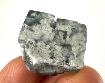May include: A green fluorite crystal with a cubic shape and a white, cloudy appearance. The crystal has a rough, natural texture.