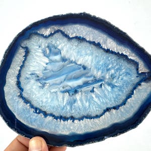 May include: A polished agate slice featuring concentric bands of deep navy blue, light blue, and translucent white. The center displays a crystalline geode formation with jagged, sparkling textures, while the outer edge is a dark, solid blue rim.