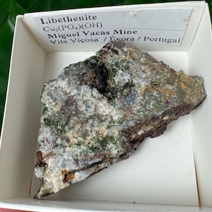 May include: A sample of Libethenite, a copper phosphate hydroxide mineral, displayed in a white box. The mineral has a rough, textured surface with shades of green, brown, and gray. The label reads "Libethenite Cu2(PO4)(OH) Miguel Vacas Mine Vila Viçosa / Evora / Portugal."