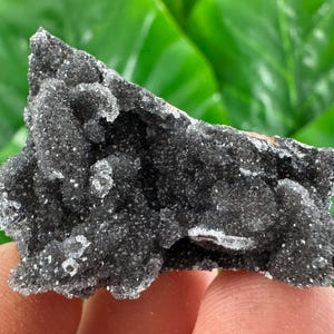 May include: A dark gray mineral specimen with a rough, textured surface. The mineral has a crystalline structure with small, sparkling inclusions. The background is a blurred green, suggesting a natural setting. The mineral is held by a person's fingers.