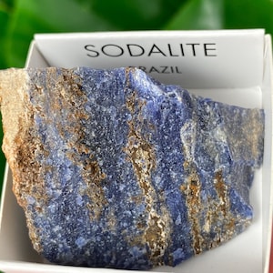 May include: A natural sodalite stone from Brazil, presented in a white box. The stone is a deep blue colour with streaks of brown and tan. The word "SODALITE" is printed on the box.