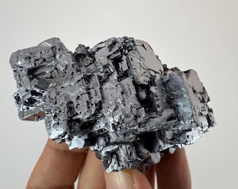 May include: A close-up of a metallic gray mineral specimen. The mineral has a rough, textured surface with a crystalline structure. The specimen is held against a plain white background.