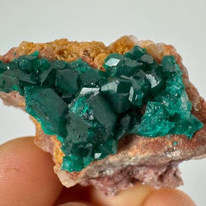 Könnte beinhalten: Ein Cluster aus leuchtend grünen Kristallen auf einer braunen und rosafarbenen Matrix. Die Kristalle haben eine scharfe, geometrische Form und sind in einem dichten Cluster angeordnet.