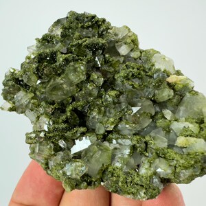 May include: A cluster of clear quartz crystals with green inclusions. The crystals are clustered together in a rough, natural formation.
