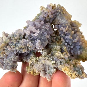 Könnte beinhalten: Nahaufnahme eines Clusters aus botryoidalem Chalcedon, einer Quarzart. Das Exemplar weist eine blasenartige, traubenähnliche Textur mit einem blassen Lavendelton auf, der durch Bereiche in Beige und Grün akzentuiert wird. Das Mineral wird in einer Hand gehalten.