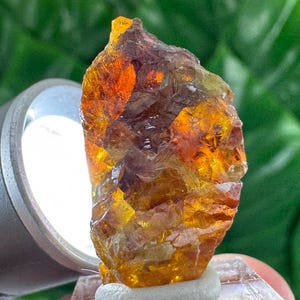 Peut inclure: Gros plan sur une pierre précieuse brute aux teintes orange, jaune et marron. Le spécimen minéral est éclairé par une lumière vive, révélant sa structure cristalline et sa beauté naturelle. La gemme est montée sur un petit socle blanc.
