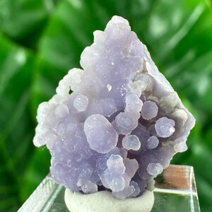 Puede incluir: Un racimo de calcedonia uva que presenta numerosas esferas pequeñas, redondeadas y translúcidas de color púrpura. La formación mineral tiene una textura botrioidal con un tono lavanda claro, mostrando una estructura cristalina natural.