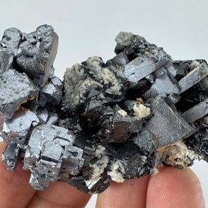 May include: A raw mineral specimen featuring metallic, dark gray cubic crystals clustered together with small patches of white, granular matrix material embedded between the sharp, reflective geometric formations of the mineral cluster.