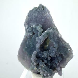 Puede incluir: Primer plano de un espécimen natural y en bruto de ágata de uva. El espécimen presenta un cúmulo de pequeñas formaciones redondas, parecidas a uvas, en tonos morados y grises. El ágata está sobre un fondo blanco.