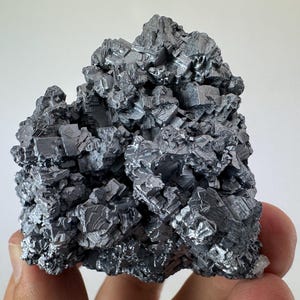 May include: A close-up of a metallic gray mineral specimen. The mineral has a crystalline structure with many small, angular, and reflective surfaces. The overall shape is irregular and textured, with a few small white crystal formations at the base.