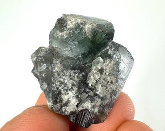 May include: A green fluorite crystal with a dark gray, almost black, exterior. The crystal has a rough, textured surface and is irregularly shaped.