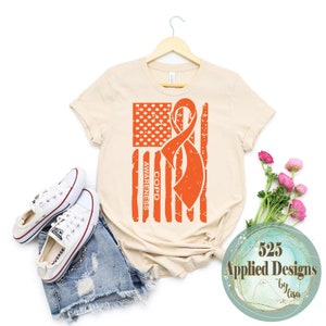 Puede incluir: Una camiseta beige con un diseño de bandera estadounidense desgastado con una cinta naranja. El texto "COPD AWARENESS" está impreso verticalmente en la bandera.