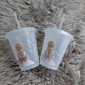 May include: Two clear plastic cups with lids and straws. Each cup features a drawing of a flower girl in a white dress with a bow. One cup says "Lily FLOWER GIRL" and the other says "Leah FLOWER GIRL".