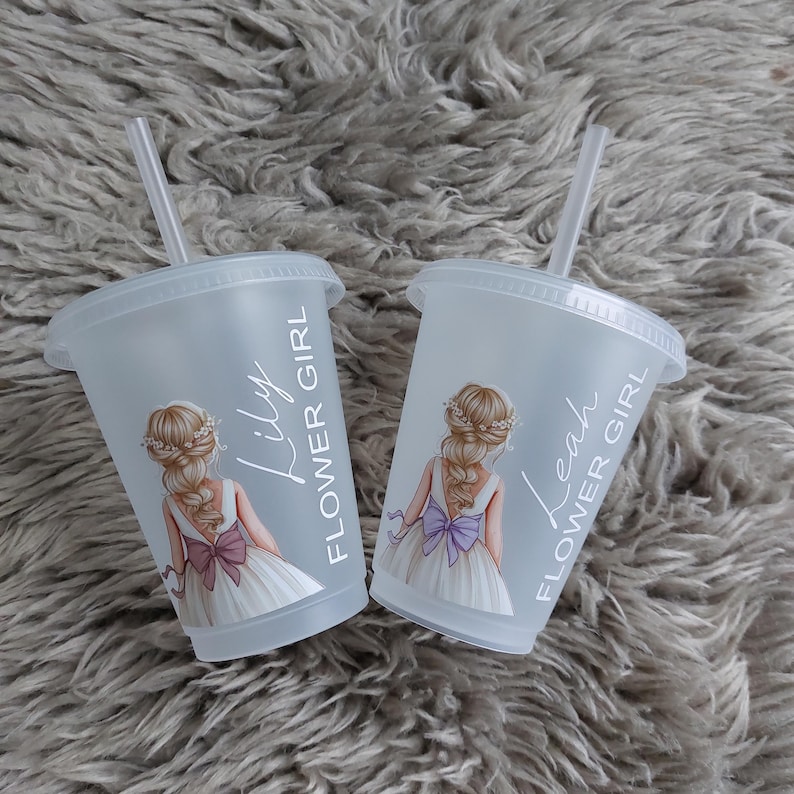 May include: Two clear plastic cups with lids and straws. Each cup features a drawing of a flower girl in a white dress with a bow. One cup says "Lily FLOWER GIRL" and the other says "Leah FLOWER GIRL".