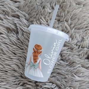 May include: A clear plastic cup with a lid and straw. The cup features a graphic of a girl in a white dress with a blue bow and the text "Olivia FLOWER GIRL". The girl has red hair and a floral crown.