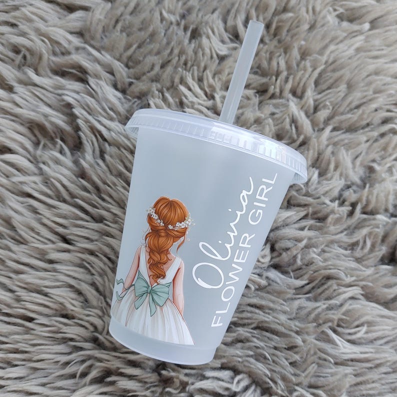 May include: A clear plastic cup with a lid and straw. The cup features a graphic of a girl in a white dress with a blue bow and the text "Olivia FLOWER GIRL". The girl has red hair and a floral crown.