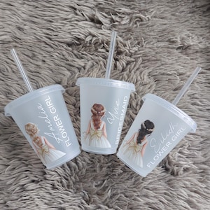 May include: Three clear plastic tumblers with lids and straws, each featuring a watercolor illustration of a girl in a white dress. Text on the cups reads "Flower Girl", "Bridesmaid", and names. The tumblers are on a gray, textured surface.