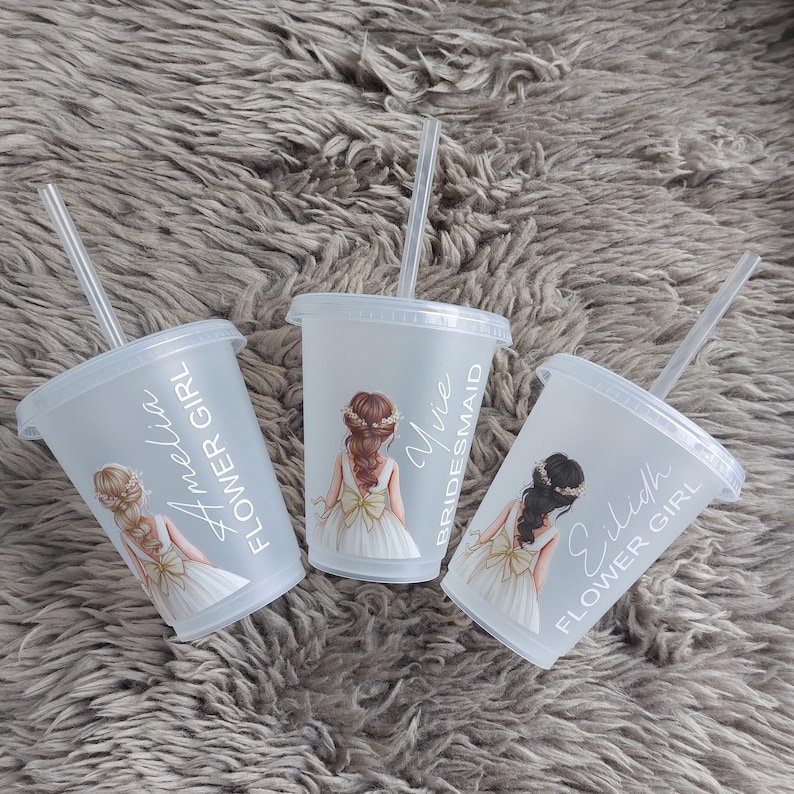 May include: Three clear plastic tumblers with lids and straws, each featuring a watercolor illustration of a girl in a white dress. Text on the cups reads "Flower Girl", "Bridesmaid", and names. The tumblers are on a gray, textured surface.