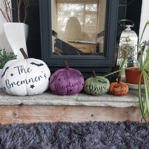May include: Four plush fabric pumpkins in white, purple, green, and orange, each featuring a brown stem, black bat and star graphics, and personalised text reading The Bremner's, The Morton's, Rhys, and Freddie in a black script font.