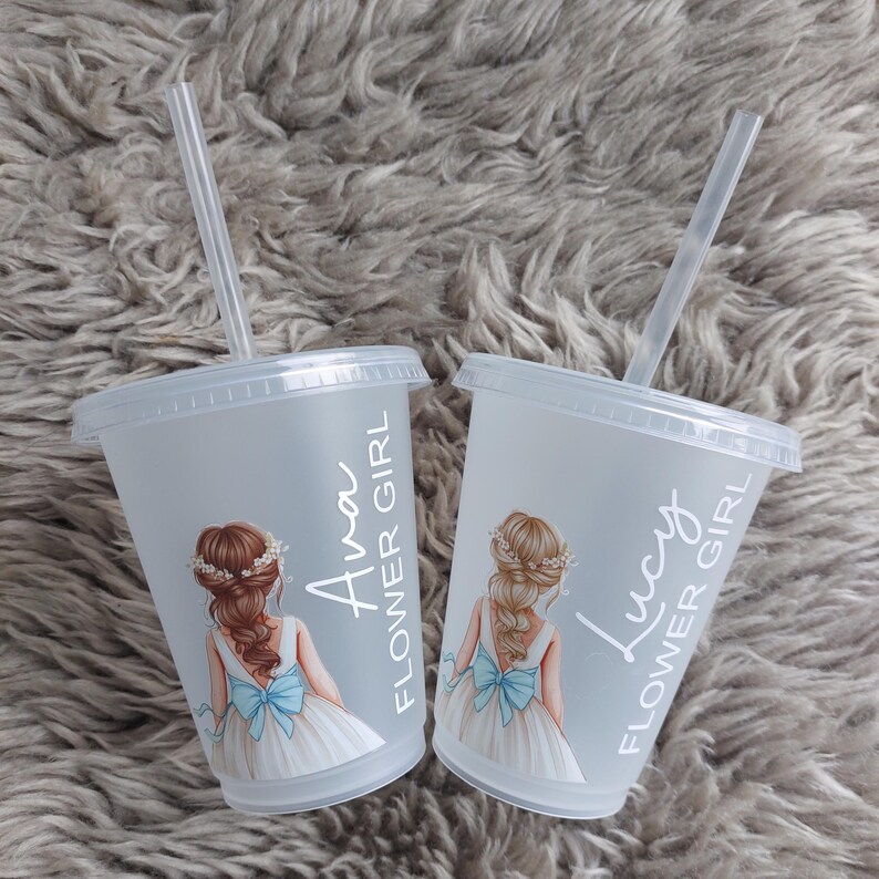 May include: Two clear plastic cups with lids and straws. Each cup has a drawing of a girl in a white dress with a blue bow. The text "Ava FLOWER GIRL" and "Lucy FLOWER GIRL" are printed on the cups.