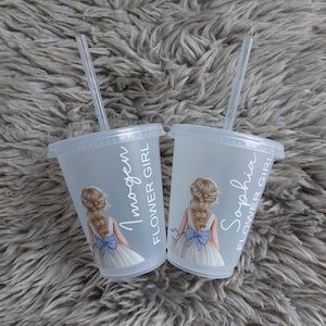 May include: Two clear plastic cups with lids and straws. Each cup features a drawing of a flower girl in a white dress with a blue bow. The names "Imogen" and "Sophia" are printed on the cups, along with the words "Flower Girl."