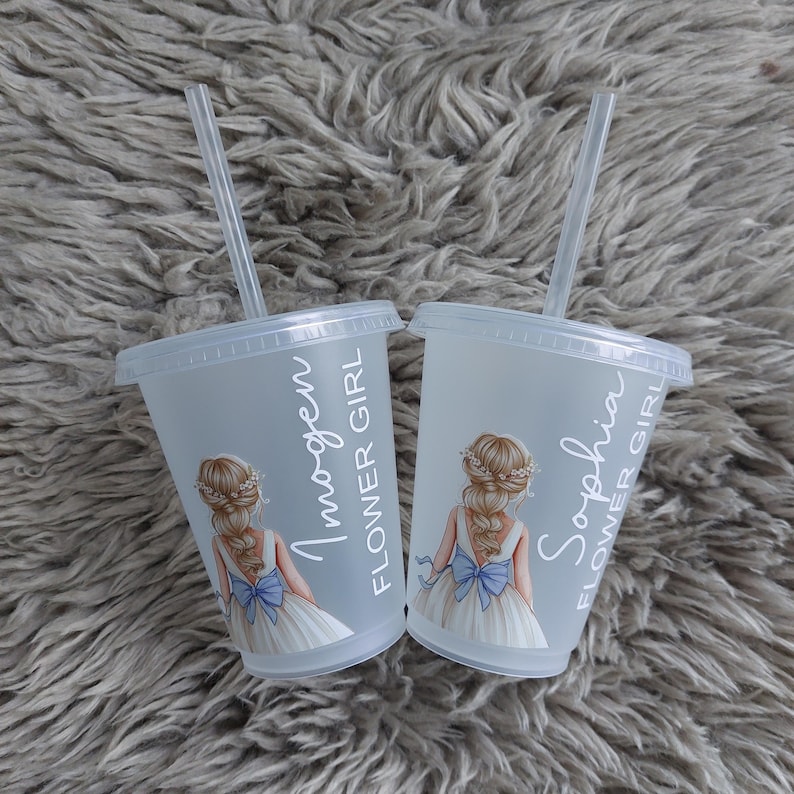 May include: Two clear plastic cups with lids and straws. Each cup features a drawing of a flower girl in a white dress with a blue bow. The names "Imogen" and "Sophia" are printed on the cups, along with the words "Flower Girl."