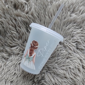 May include: A clear plastic tumbler with a lid and straw. The tumbler features a graphic of a girl in a white dress with a green bow. The text "Piper Confetti Girl" is printed on the side. The tumbler is on a gray, fluffy surface.