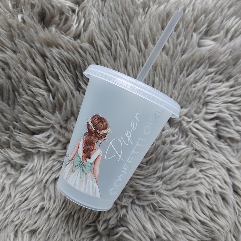 May include: A clear plastic tumbler with a lid and straw. The tumbler features a graphic of a girl in a white dress with a green bow. The text "Piper Confetti Girl" is printed on the side. The tumbler is on a gray, fluffy surface.