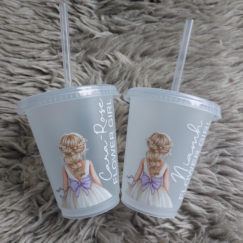 May include: Two frosted plastic tumblers with lids and straws. Each cup features a drawing of a girl in a white dress with a purple bow. Text on the cups reads "Cara-Rose Flower Girl" and "Niamh Flower Girl".