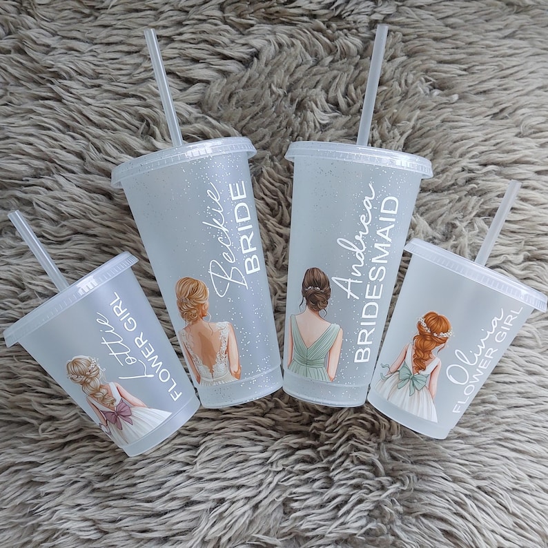 May include: Four clear plastic tumblers with lids and straws. Each tumbler has a different name and role: "Beckie Bride", "Andrea Bridesmaid", "Olivia Flower Girl", and "Lottie Flower Girl". The tumblers have illustrations of women in wedding attire.