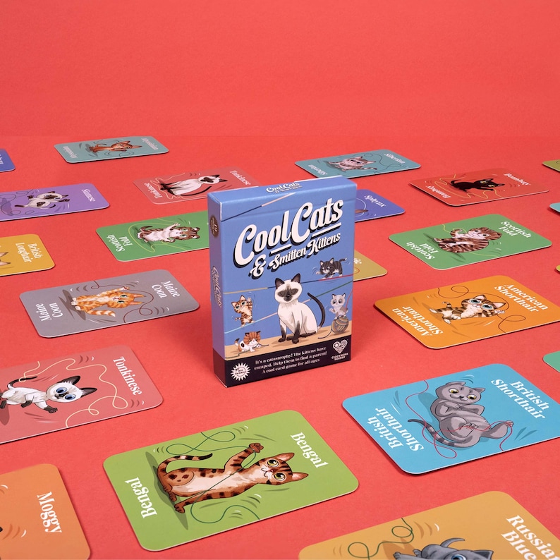 May include: A card game box titled "Cool Cats & Smitten Kittens" with illustrations of various cat breeds. The game cards are scattered around the box, each featuring a different cat breed with its name. The background is a solid red.
